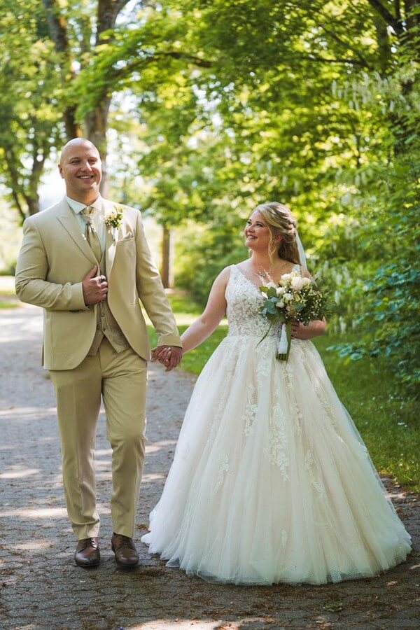 Curvy Braut in Prinzessinnenkleid mit langer Schleppe (Größe 46) und Bräutigam im beigen Anzug mit Weste und Plastron – Hochzeitsmode für echte Paare bei Biancas Brautmoden