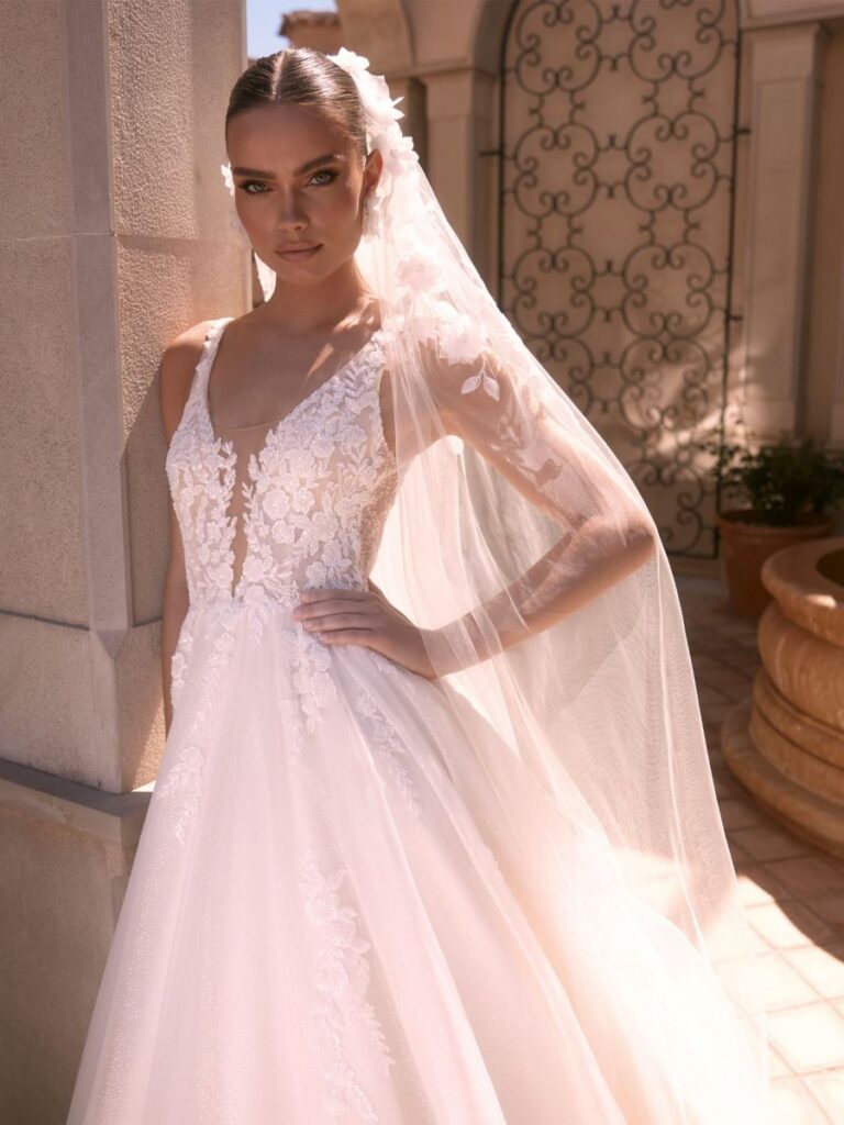 Brautkleid mit tiefem V-Ausschnitt, Spitzenoberteil, Glitzertüllrock und langem Schleier mit Blüten von Bianco Evento 2026