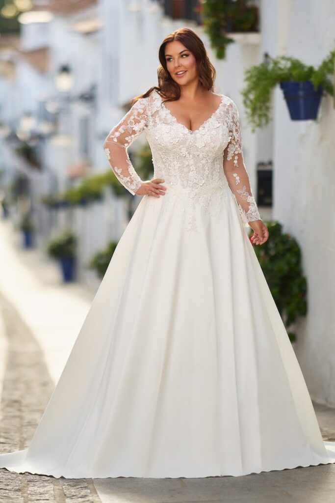 Edles Curvy Brautkleid 2026 in A-Linie mit transparenter Spitzen-Corsage, langen Ärmeln und romantischen Blütenapplikationen – erhältlich bei Bianca’s Brautmoden.